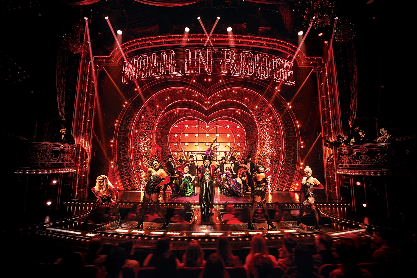 Scene from Moulin Rouge! The Musical on Broadway, featuring dancers in extravagant costumes on a lavish red and gold stage with a heart-shaped illuminated backdrop and “MOULIN ROUGE” marquee.