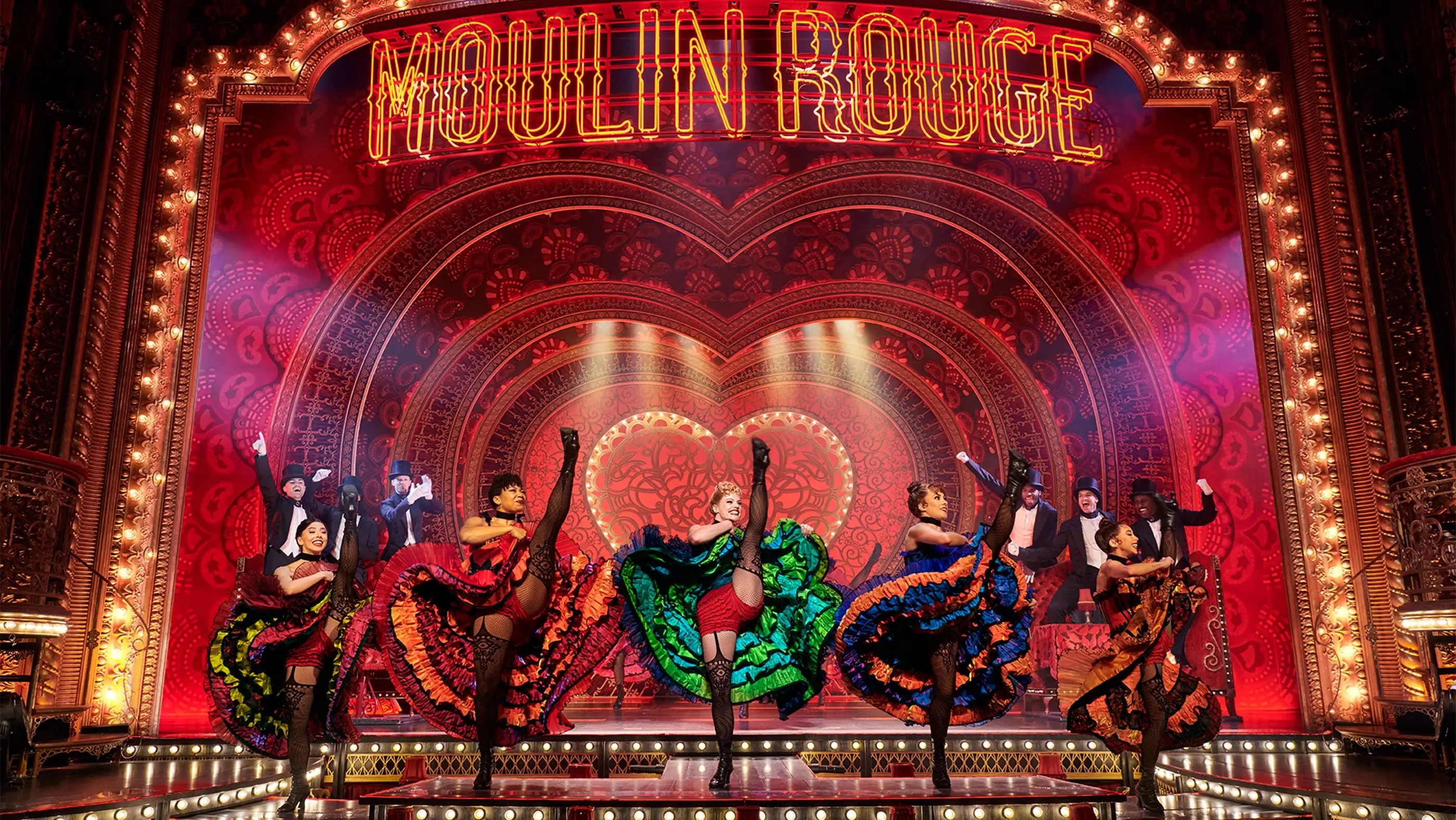 Scene from Moulin Rouge! The Musical on the West End, with dancers in colorful costumes performing can-can kicks on an ornate stage featuring a neon “MOULIN ROUGE” sign and heart-shaped backdrop.