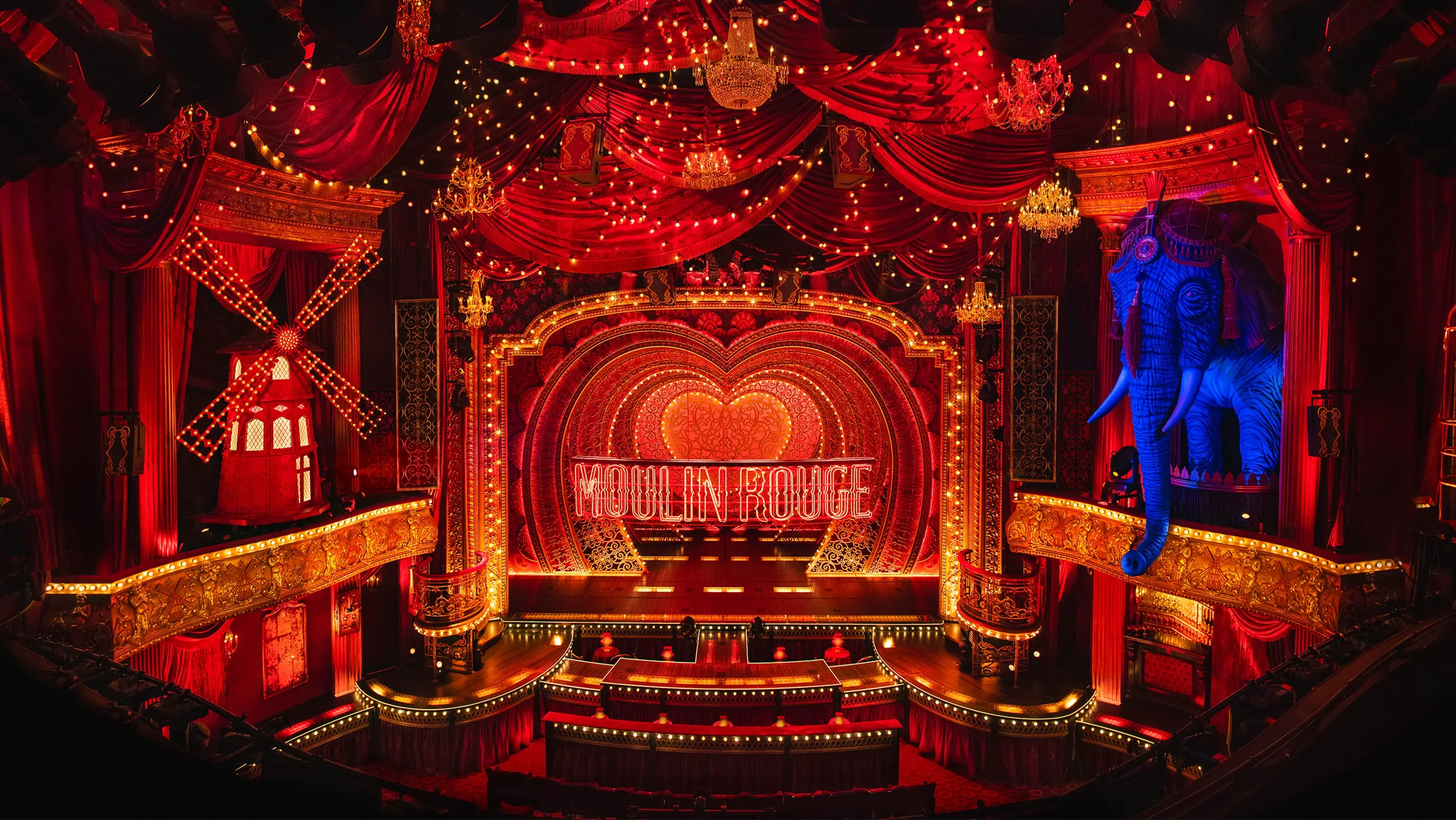 A stage set from Moulin Rouge! The Musical, featuring a richly decorated interior with red velvet drapes, ornate chandeliers, and a glowing 