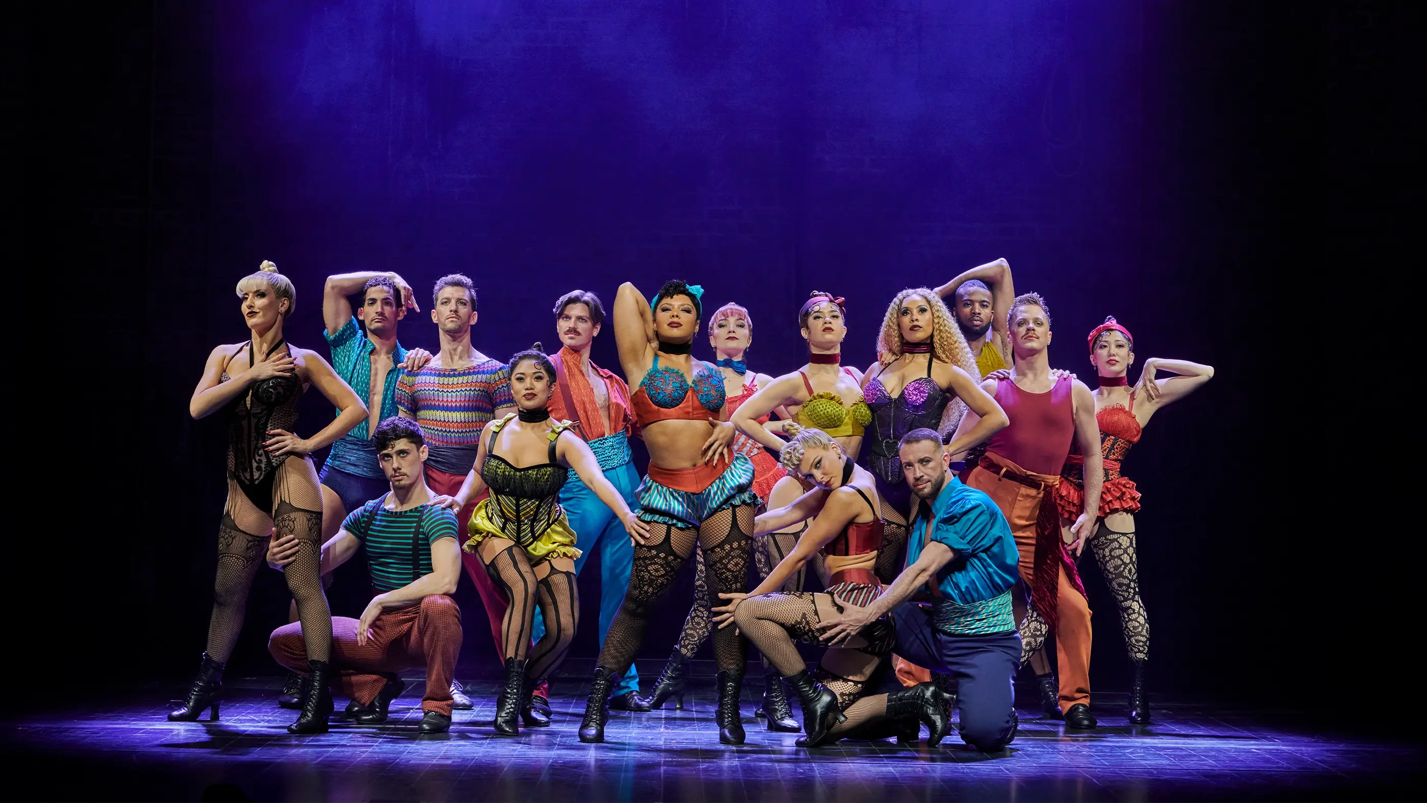 Scene from Moulin Rouge! The Musical World Tour during “Backstage Romance,” featuring performers in vibrant theatrical costumes posed dramatically under stage lighting.