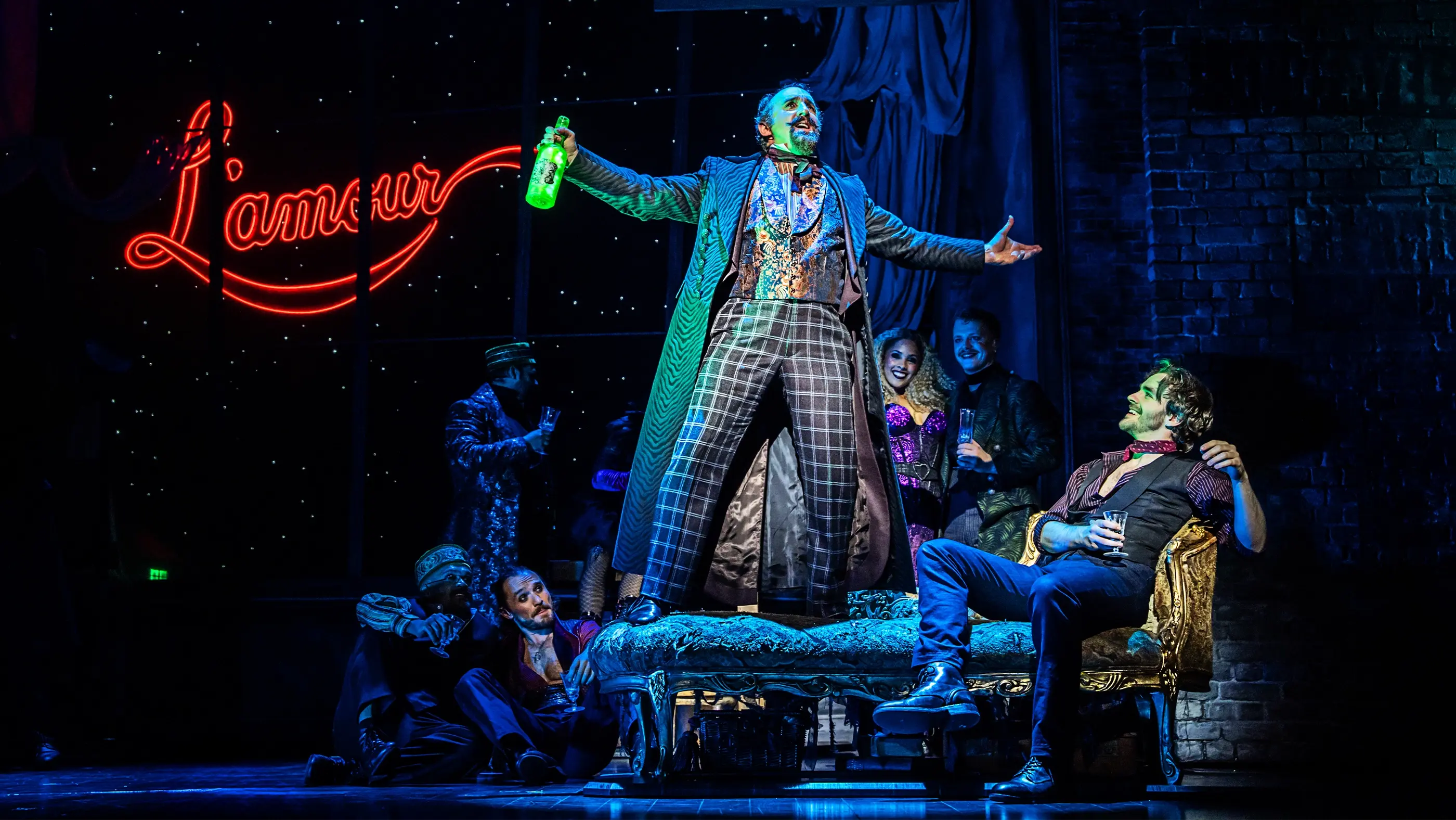 Scene from Moulin Rouge! The Musical World Tour featuring several actors on stage. Zidler stands on a chaise lounge holding a green bottle of Absinthe and gesturing dramatically, while others are seated or standing nearby in elaborate costumes. A red neon sign reading 