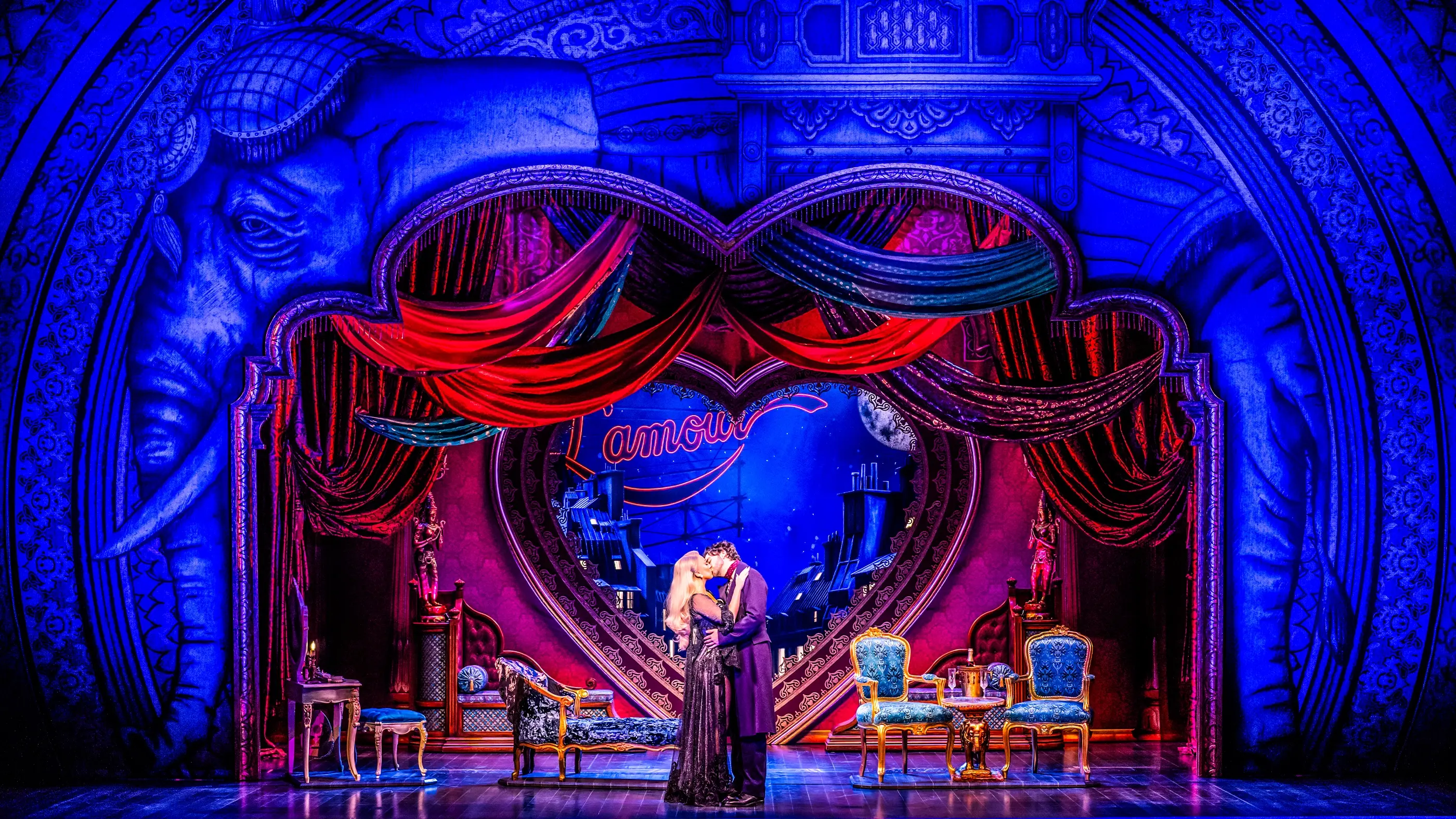 Romantic stage scene from Moulin Rouge! The Musical World Tour. Christian and Satine stand center stage in an intimate moment. The backdrop features a large heart, red drapery, and the word “l’amour” in cursive. Ornate furniture including chairs and a chaise lounge decorate the set. A night sky with a full moon and Parisian rooftops completes the atmospheric, love-themed setting.