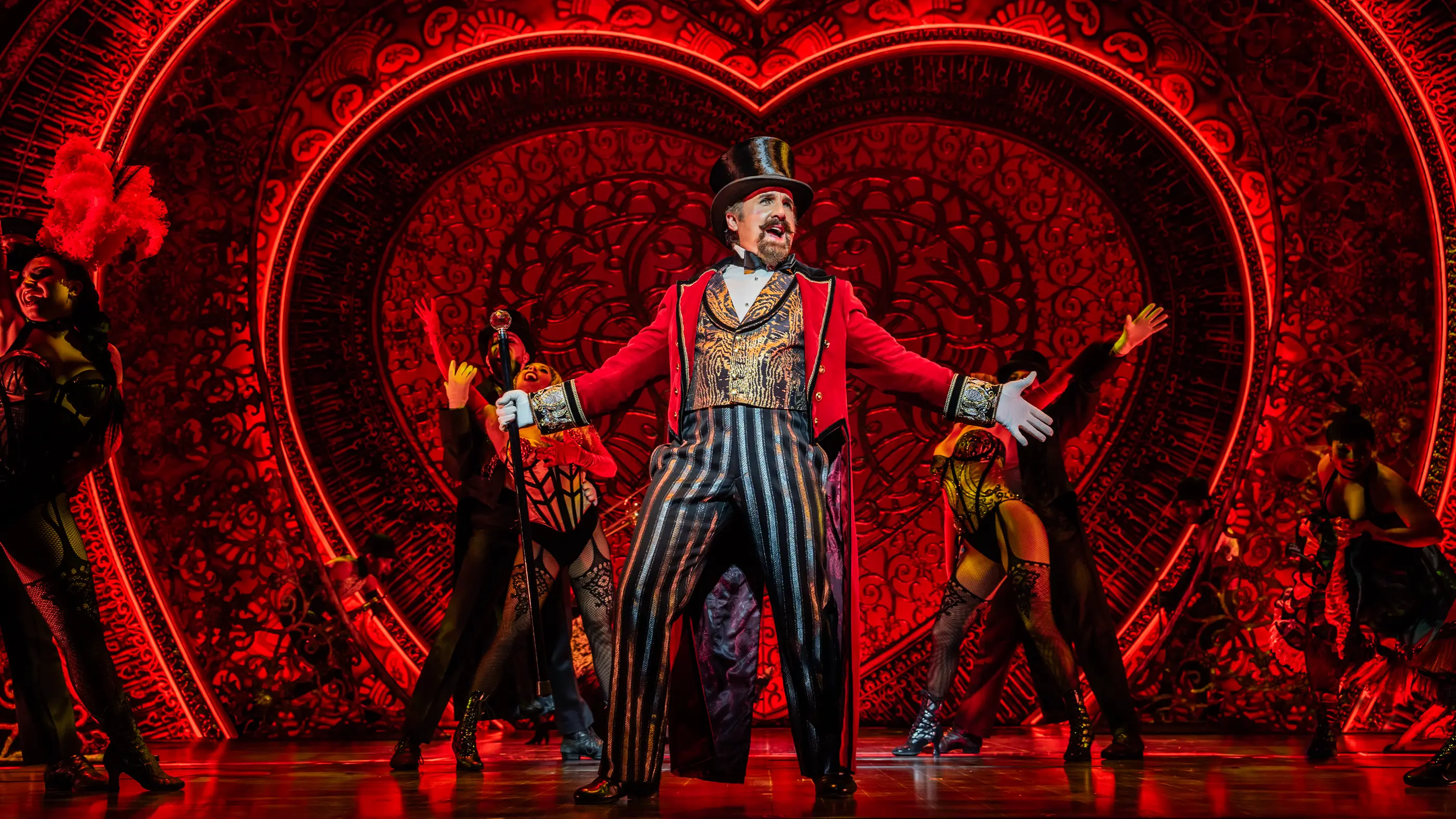 A vibrant stage scene from the Moulin Rouge! The Musical World Tour. The backdrop features a large illuminated heart with intricate designs and red lighting. Zidler stands centre stage in formal attire holding a cane, appearing to lead the performance. The scene is lively and rich in red tones.