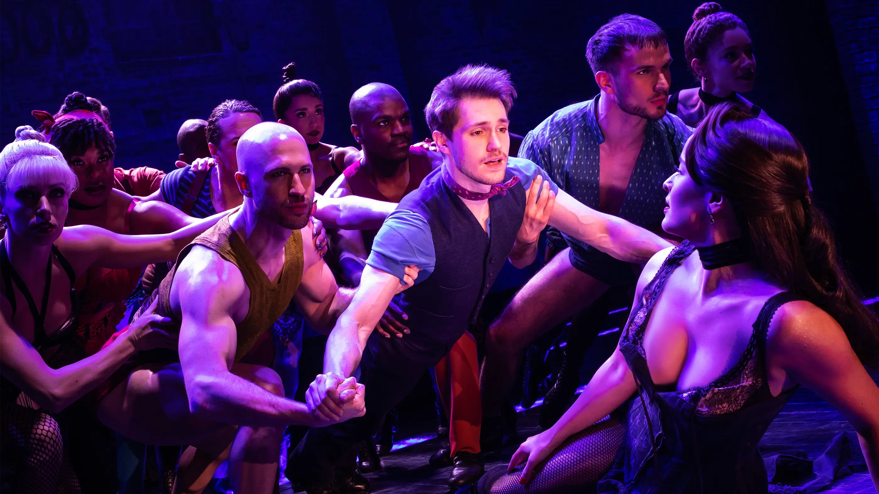 Scene from Moulin Rouge! The Musical (North American Tour), during “Backstage Romance.” Christian leans toward Satine, who is leaning back in a black corset and fishnet stockings. The stage is bathed in dramatic purple lighting, with other performers in dynamic poses surrounding them.