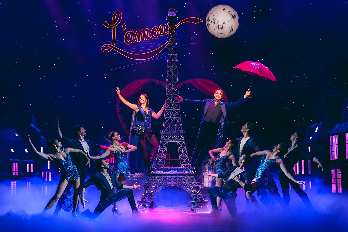 Scene from Moulin Rouge! The Musical Netherlands during “Elephant Love Medley,” with two main characters beside a central Eiffel Tower structure, one holding a red umbrella, under starry lights and a glowing “L’amour” sign.