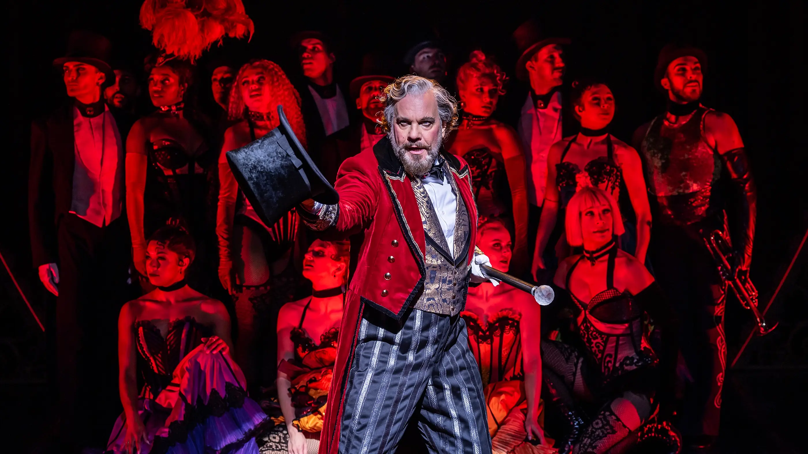 A theatrical scene from Moulin Rouge! The Musical (Netherlands production). Center stage is the character Harold Zidler, wearing a red velvet tailcoat with gold buttons, a gold-and-black patterned vest, striped pants, and holding a black top hat and cane. He is surrounded by performers in elaborate cabaret-style costumes—corsets, fishnets, top hats, and feathers—set against a dark background with dramatic red lighting.