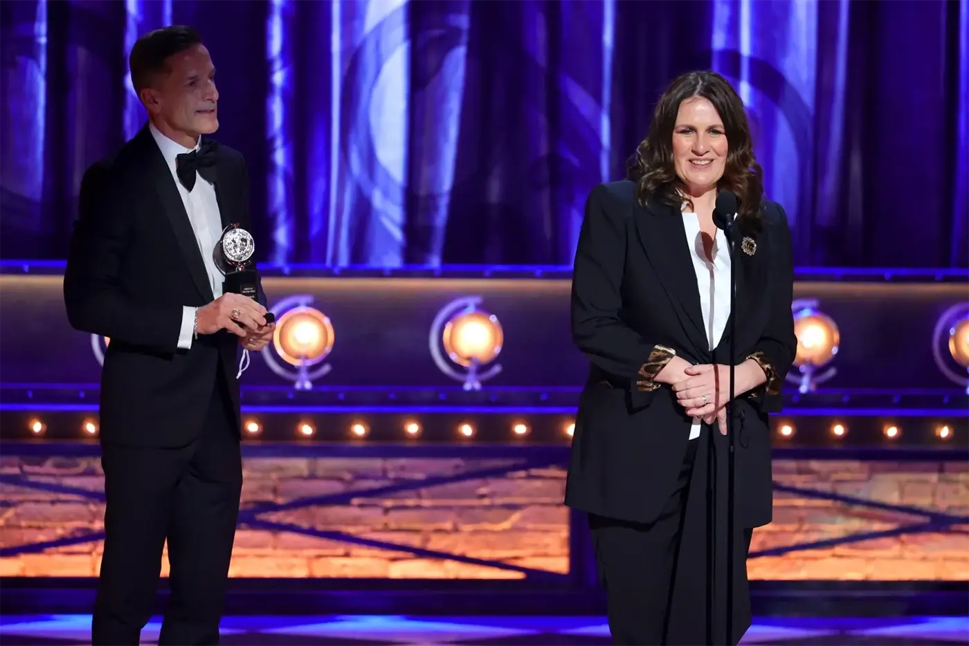 Producers of Moulin Rouge! The Musical on stage at the Tony Awards, with one holding a trophy and the other speaking into a microphone against a backdrop of blue curtains and stage lights.