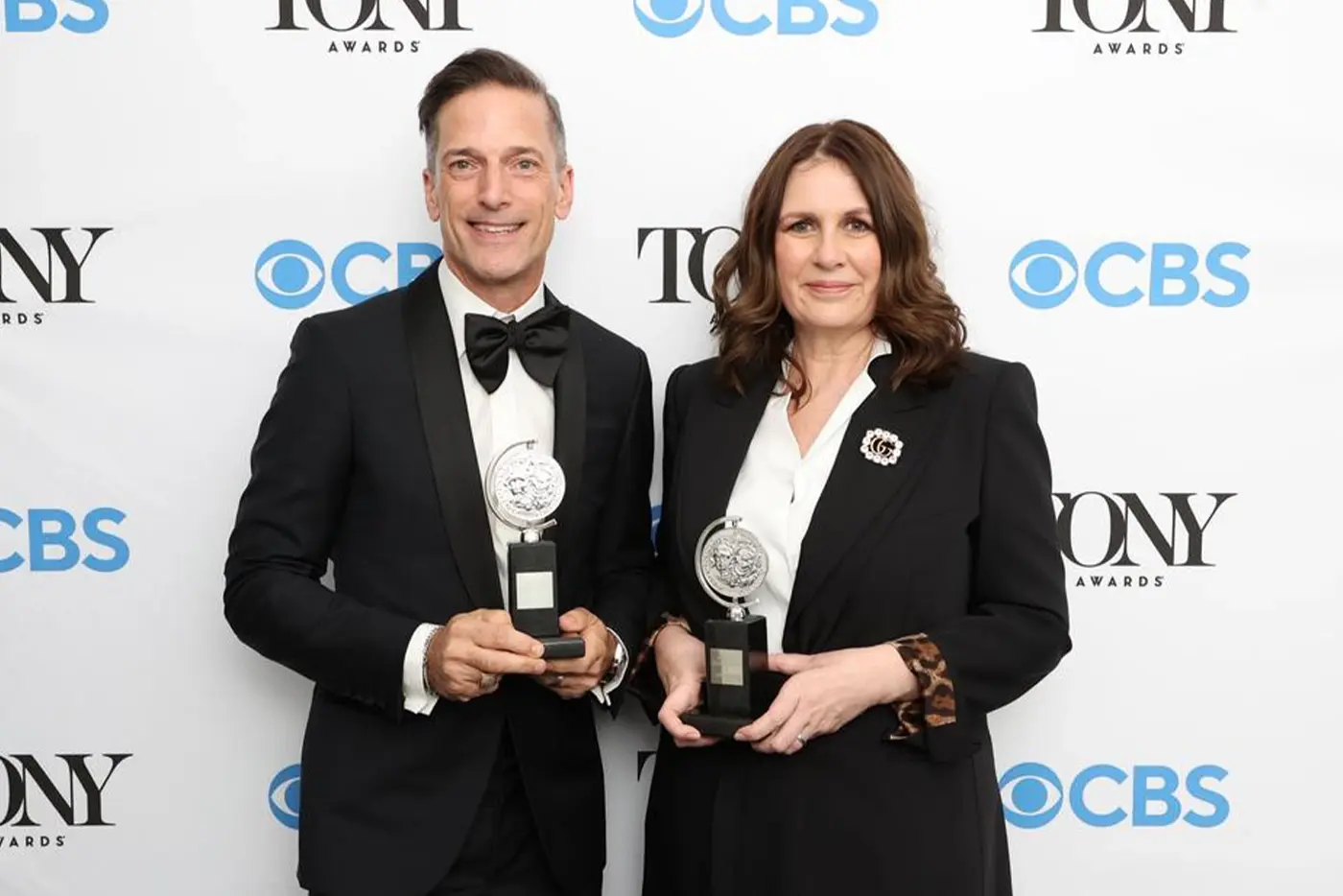 Carmen Pavlovic and Bill Damaschke in formal attire holding Tony Awards, standing in front of a backdrop featuring repeated Tony Awards and CBS logos; one wears a black tuxedo with a bow tie, the other a black suit with a white shirt.