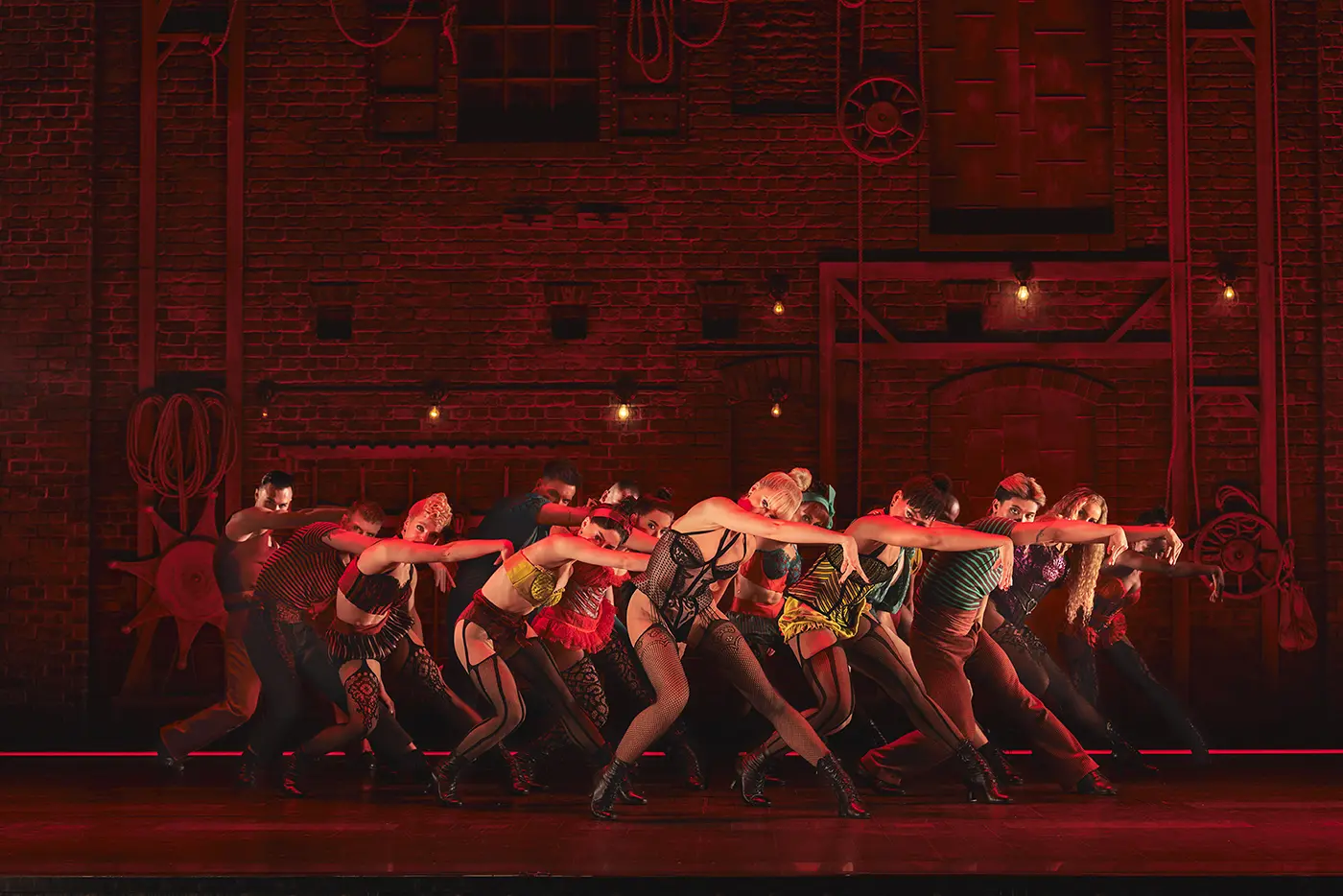 Scene from Moulin Rouge! The Musical (West End) during “Backstage Romance.” A group of dancers performs in front of a red brick wall with ropes hanging down, evoking a backstage atmosphere. The dancers wear colorful, cabaret-style costumes and strike dynamic poses with arms extended forward, capturing the energy of the number.