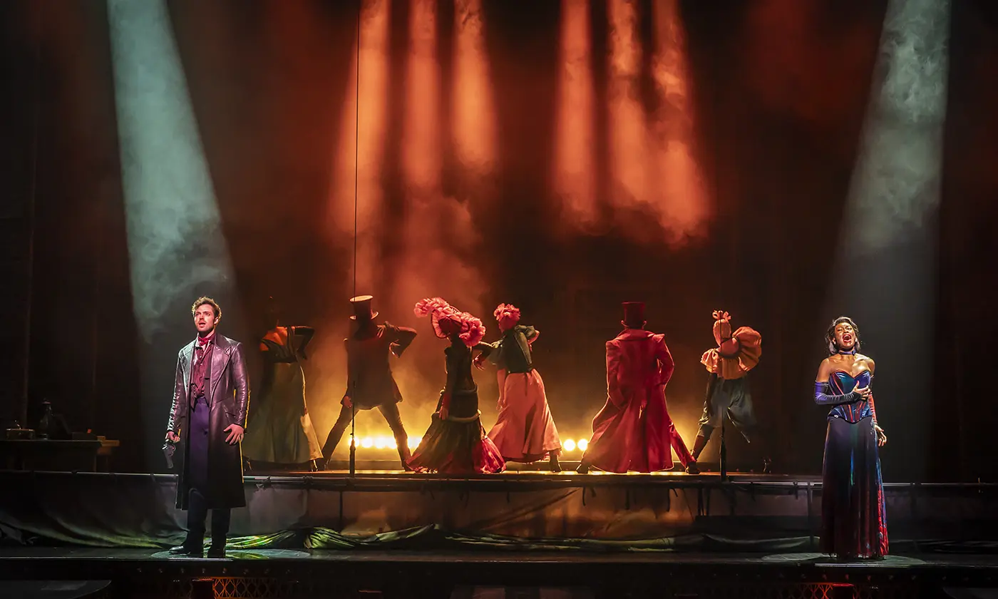 A theatrical scene from Moulin Rouge! The Musical West End featuring eight performers on stage. Chrisian is on the far left and Satine is on the far right. They each have a spotlight shining on them. In the background, the other performers face backwards, wearing elaborate costumes with a historical flair. The stage is lit with dramatic orange and blue lighting, and the background is filled with smoke or fog, adding to the intensity and atmosphere of the performance.