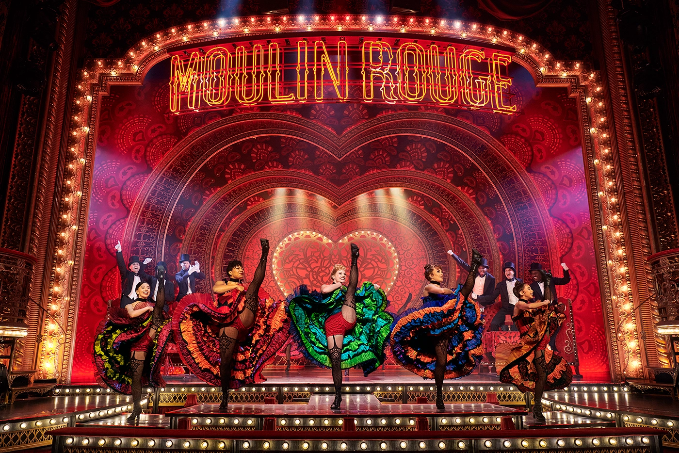 Scene from Moulin Rouge! The Musical on the West End, with dancers in colorful costumes performing can-can kicks on an ornate stage featuring a neon “MOULIN ROUGE” sign and heart-shaped backdrop.