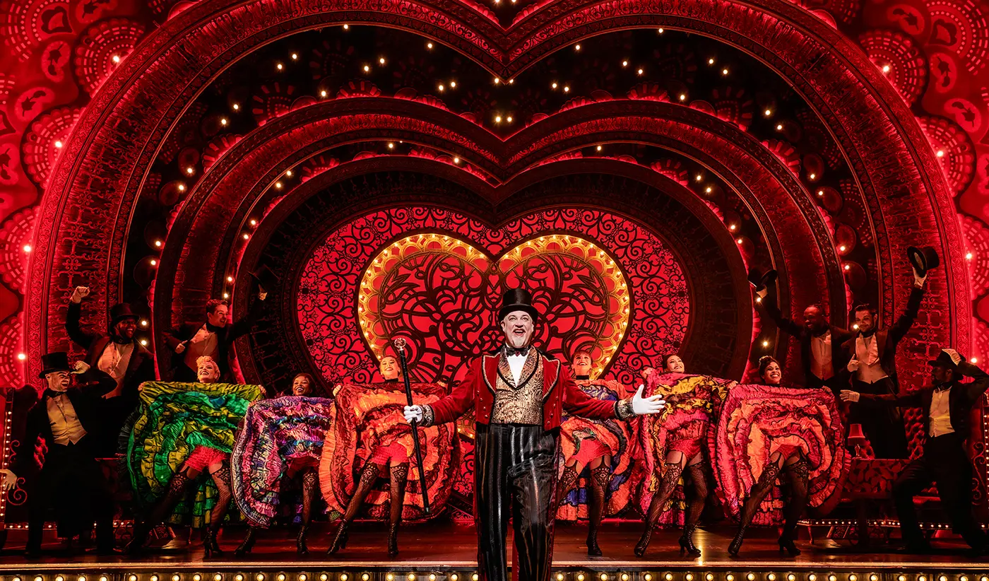 A vibrant stage scene from Moulin Rouge! The Musical on the West End. The backdrop features a large illuminated heart with intricate designs and red lighting. Multiple performers are on stage, including dancers in colorful costumes with ruffled skirts and fishnet stockings. A central figure in formal attire holds a cane, appearing to lead the performance. The scene is lively and rich in red tones.