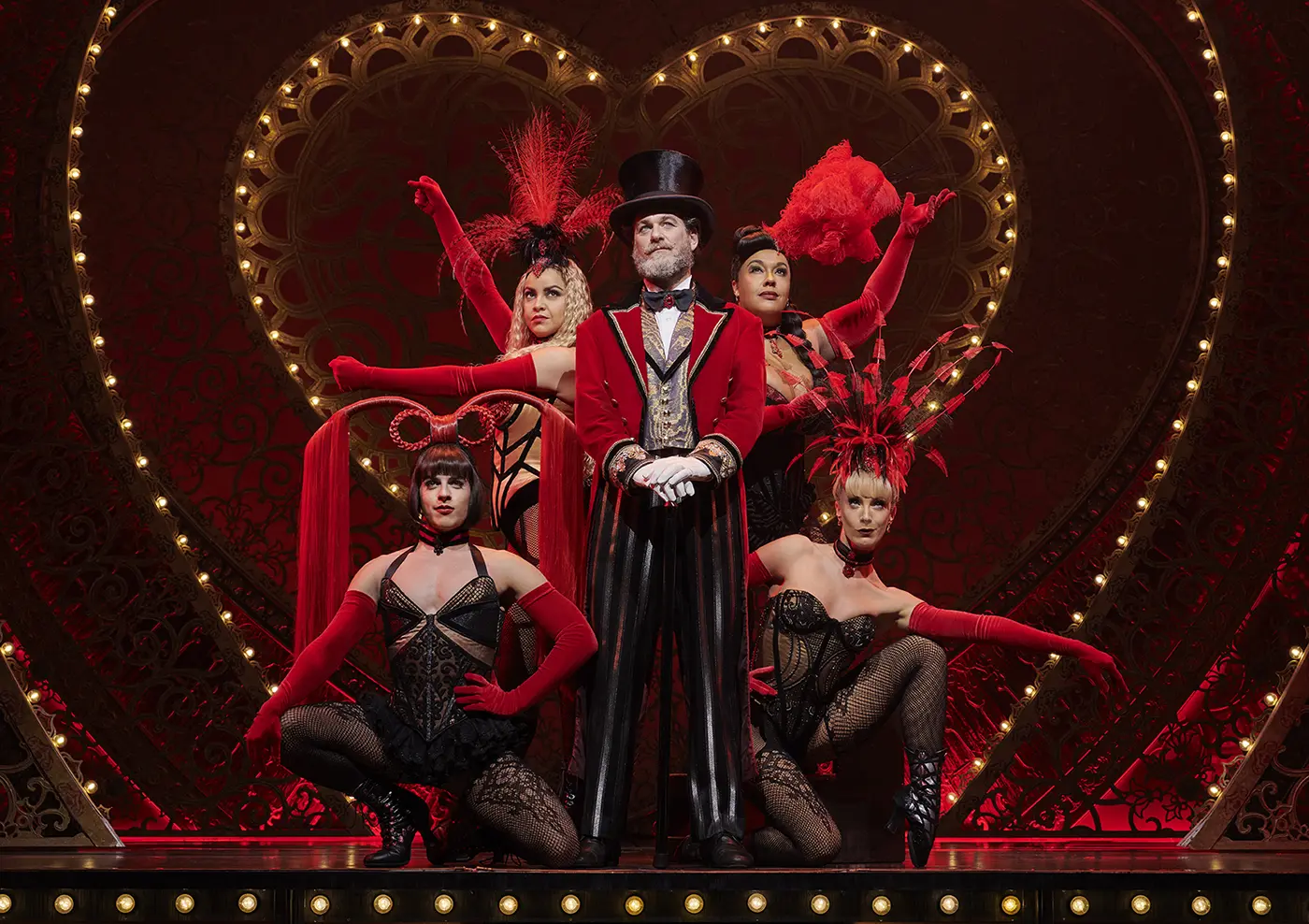 Scene from Moulin Rouge! The Musical (Broadway) featuring Harold Zidler center stage. He wears a red and gold ornate jacket, black and white striped pants, a top hat, and holds a cane. Surrounding him are the four Lady Ms, dressed in corsets, fishnet stockings, gloves, and feathered headpieces. The background is richly decorated with red lighting and intricate patterns.
