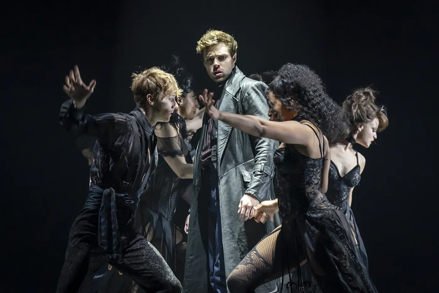 A theatrical scene from Moulin Rouge! The Musical West End. Five performers are on stage, with the Christian in the centre wearing a long grey coat. Four others in dark, dramatic costumes surround him, extending their arms in expressive poses. The dark background highlights the performers and their movements.