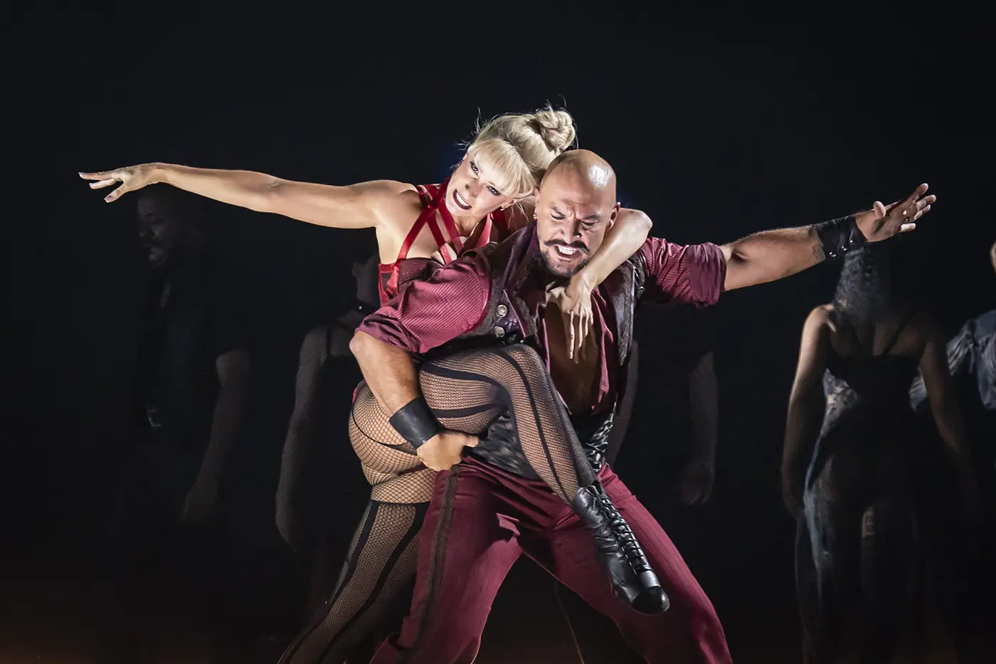 A theatrical scene from Moulin Rouge! The Musical West End. Nini is wearing a red corset and wrapped around Santiago's back while he drags her in a dramatic dance move. Both their arms are extended. The background is dimly lit.