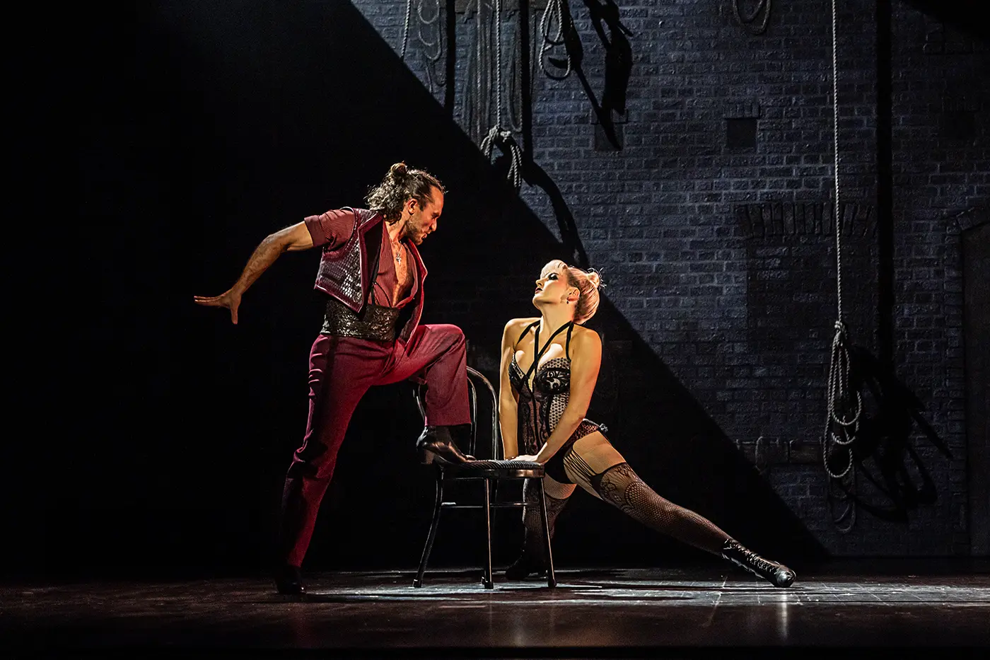 A scene from Moulin Rouge! The Musical World Tour between Santiago and Nini. Santiago stands on the left with one foot on a chair, wearing a red outfit with an open shirt and black boots. Nini sits on a chair to the right, leaning forward with one leg extended, dressed in a black corseted costume with fishnet stockings and high-heeled boots. The backdrop is a brick wall with hanging ropes, evoking a backstage setting.