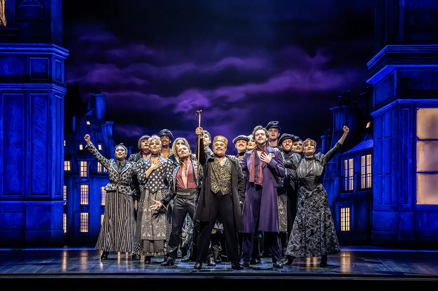 A theatrical scene from Moulin Rouge! The Musical World Tour. A group of performers stands on stage against a backdrop of a dark, cloudy sky and buildings with glowing windows. Toulouse-Lautrec stands in the centre holding up a staff, and all are dressed in period costumes, contributing to the dramatic atmosphere.