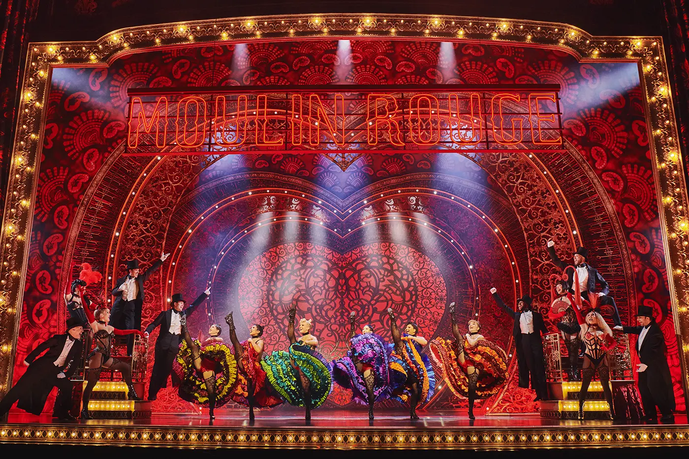 Scene from Moulin Rouge! The Musical World Tour, with dancers in colorful costumes performing can-can kicks on an ornate stage featuring a neon “MOULIN ROUGE” sign and heart-shaped backdrop.