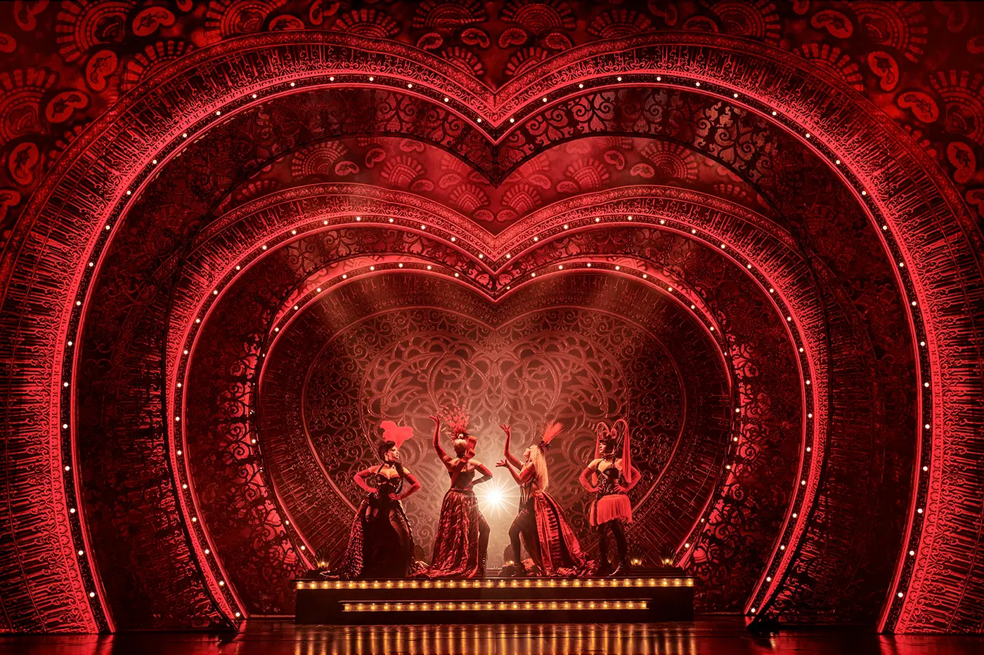 Scene from Moulin Rouge! The Musical World Tour featuring the Lady M’s in red and black burlesque costumes with feathered headpieces, posed in front of an ornate heart-shaped stage backdrop.