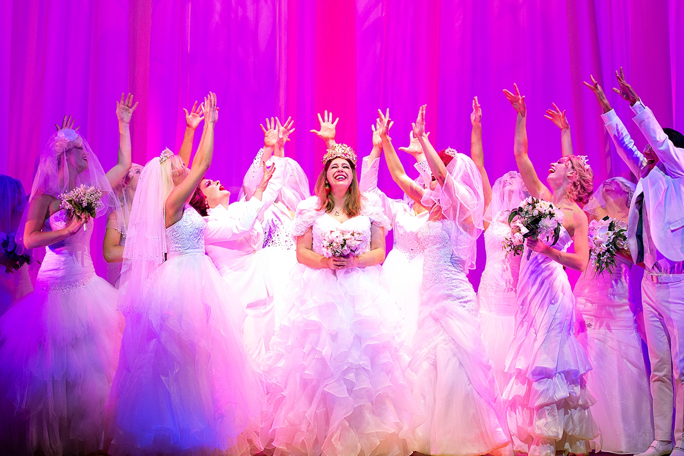 Scene from Muriel’s Wedding The Musical in Australia, featuring performers in bridal attire celebrating around Muriel holding a bouquet, under vibrant pink and purple lighting.