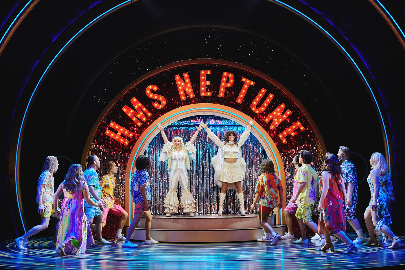 Scene from Muriel’s Wedding The Musical in Leicester, featuring two central performers in white costumes on an elevated platform under an “H.M.S NEPTUNE” sign, surrounded by dancers in tropical-themed outfits and sparkling streamers.