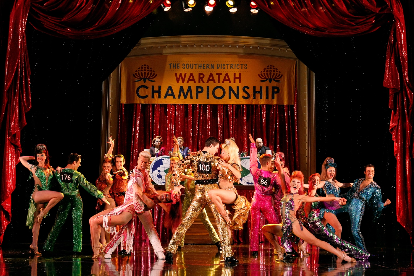 Scene from Strictly Ballroom The Musical Australia, with dancers in glittering costumes performing under a banner reading “The Southern Districts Waratah Championship,” featuring a central couple with competition numbers 100.