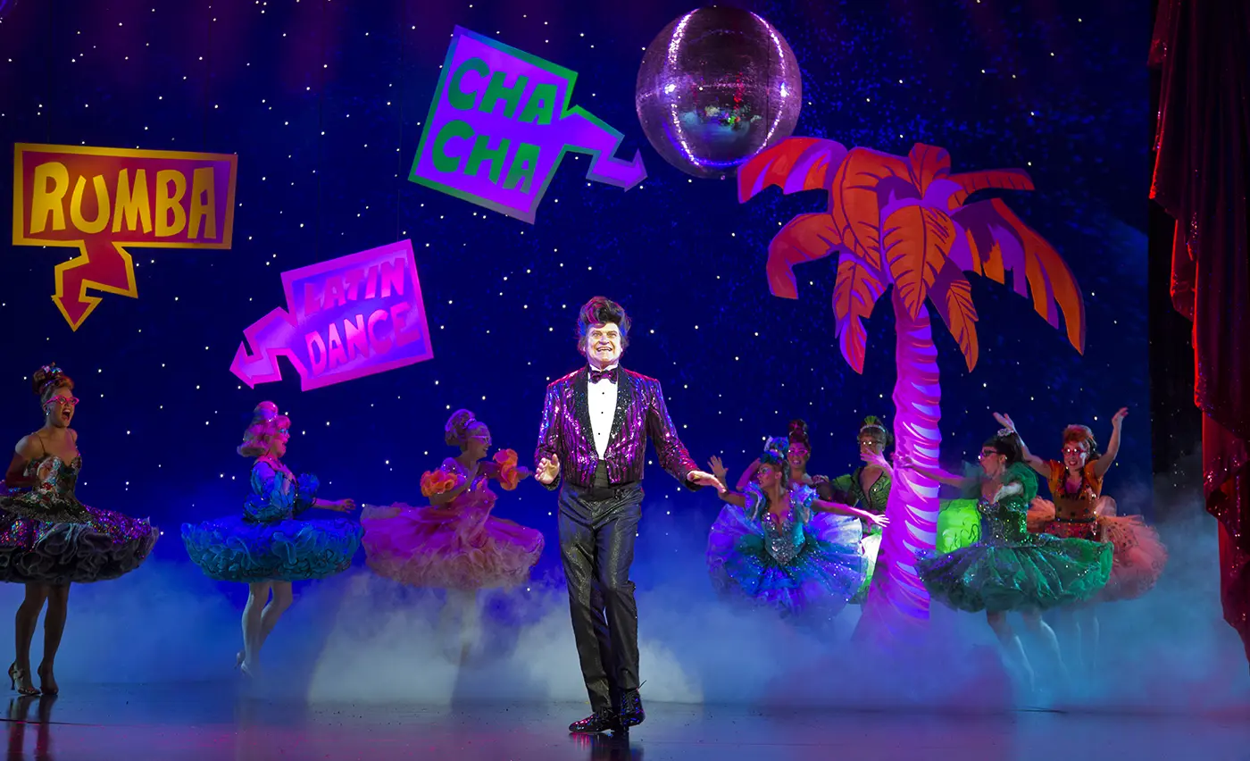 Scotts dad Doug Hastings dancing in a purple sequin jacket with dancers in the background under signs Rumba, Latin and Cha Cha.