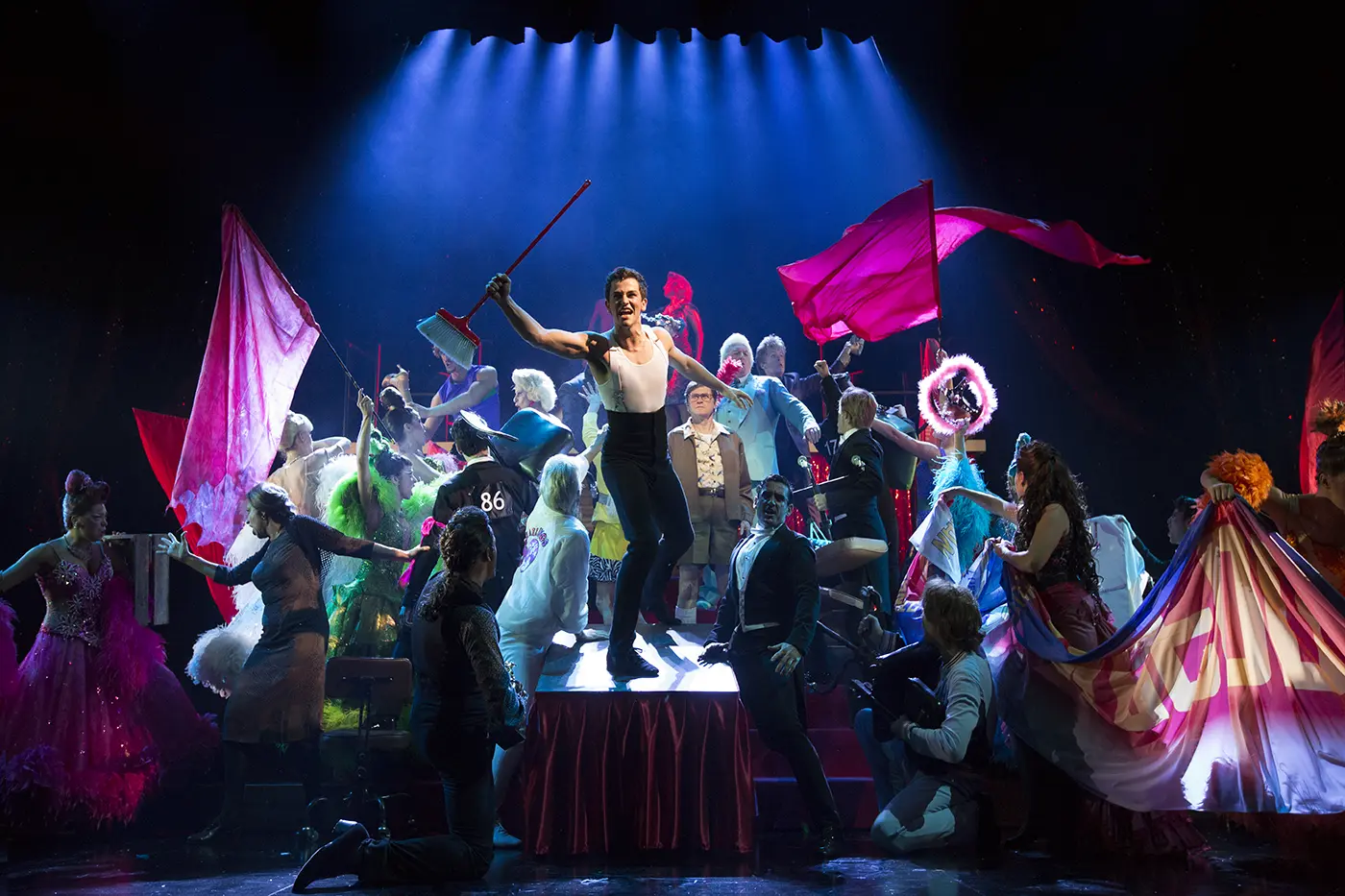 Scott holding a broom up staning on top of a stage while the cast around are in all different positions and holding different things like pink flags, and banners