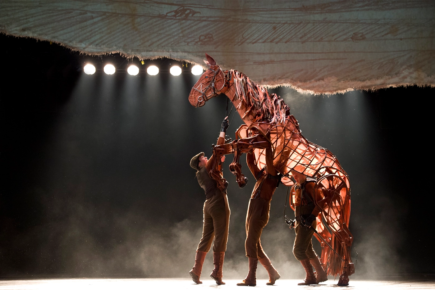 Scene from War Horse Australia featuring a life-sized horse puppet operated by three performers in period military uniforms under dramatic spotlighting.