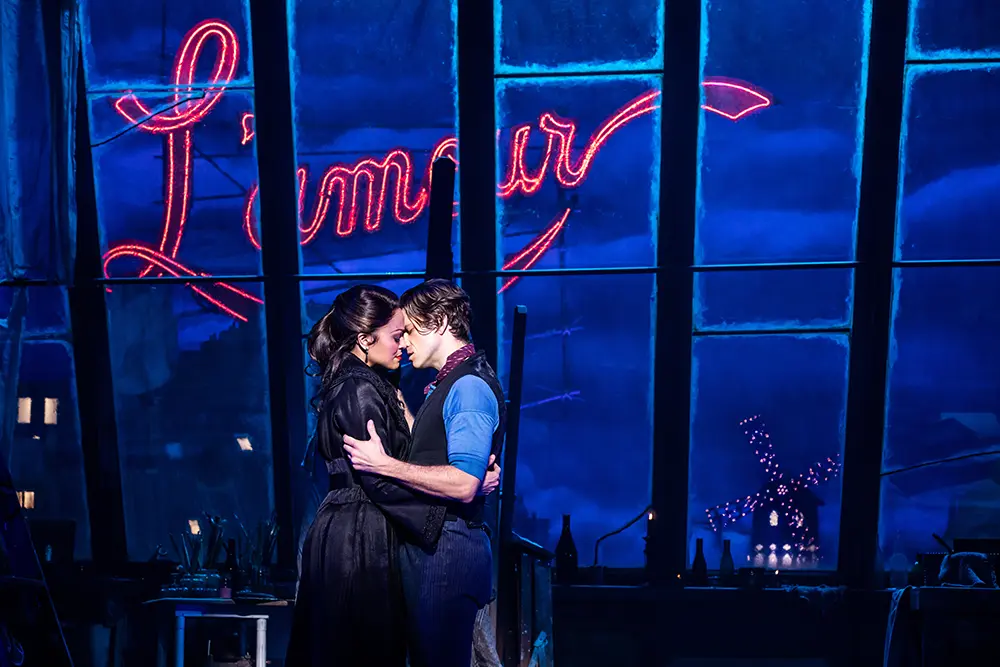 Scene from Moulin Rouge! The Musical in “Come What May,” with Christian and Satine embracing under blue lighting, framed by large windows and a glowing “L’amour” sign.