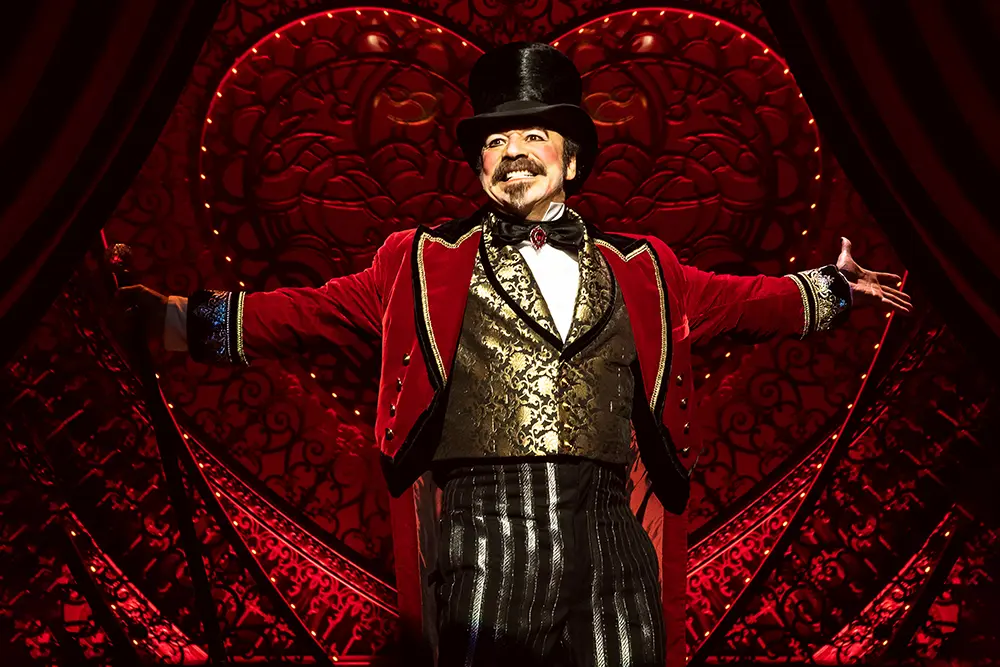Scene from Moulin Rouge! The Musical (Broadway cast) featuring Danny Burstein as Harold Zidler spreading his arms wide with a teethy grin standing in front of the heart-shaped set.